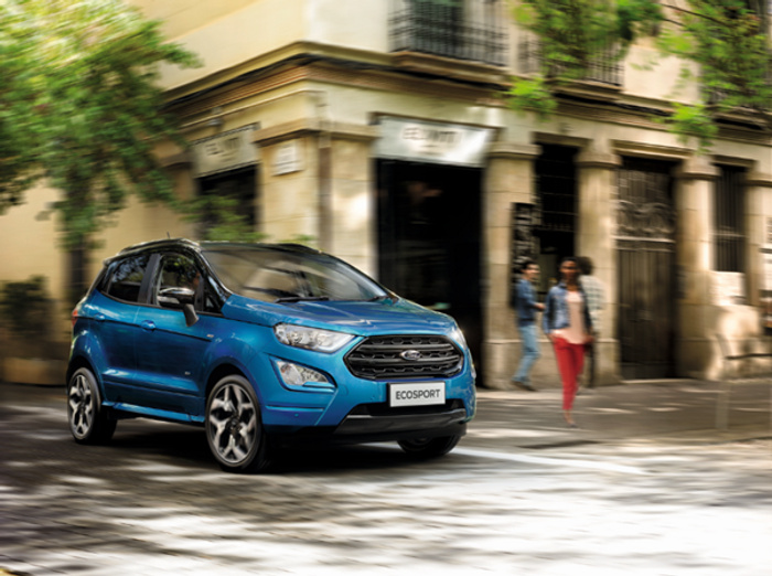 Blue Ford EcoSport compact SUV driving on a sunlit urban street with blurred background featuring an old building and pedestrians walking by, highlighting a new car model suitable for city driving, ideal for buyers interested in used cars or new cars.