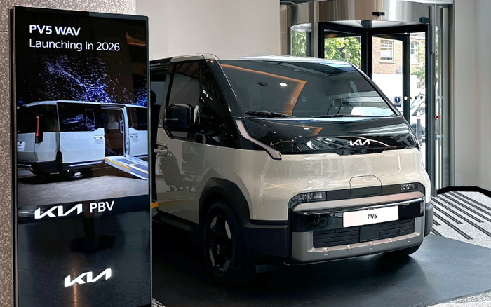 Kia PV5 WAV electric people mover vehicle displayed indoors next to a digital sign announcing 'PV5 WAV Launching in 2026' and showing a side view of the vehicle with a wheelchair ramp. The modern compact van is white with black detailing, branded with the Kia logo. This image highlights innovative new cars concepts, relevant for used cars and new cars markets.