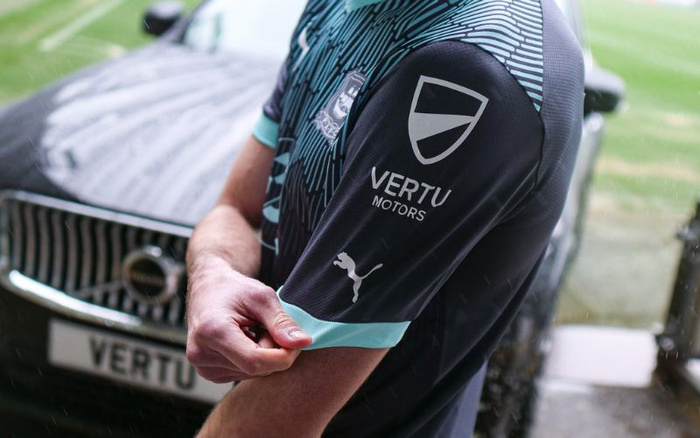 Close-up of a person wearing a black and turquoise sports jersey with logos including Vertu Motors and Puma on the sleeve, standing in front of a car with a visible grille and a license plate reading 'VERTU', representing the Vertu Motors brand associated with new cars and used cars.