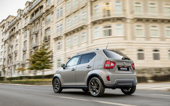 Silver Suzuki Ignis compact car driving on city street with large historic building in the background, showcasing a modern used car option suitable for urban environments, ideal for buyers interested in new cars or used cars.