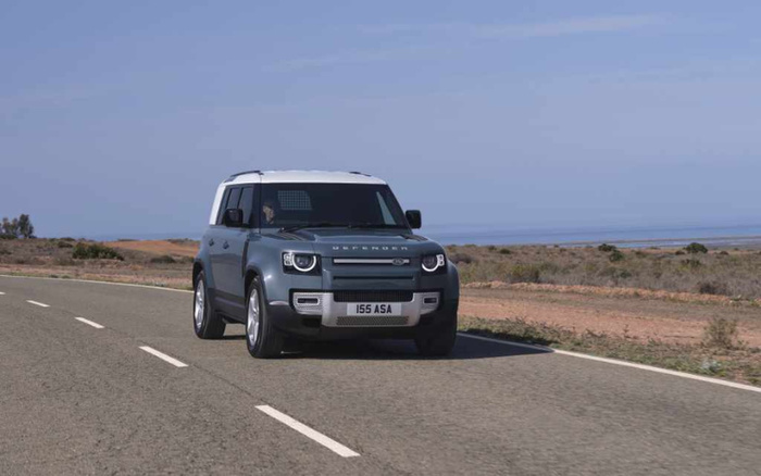 Blue Land Rover Defender driving on a deserted road with dry shrubland and coastline in the background, showing license plate number 155 ASA, representing quality new cars and used cars in excellent condition.