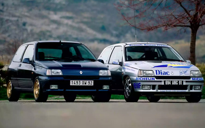 Two Renault Clio cars parked side by side on a road with a scenic blurred background. The car on the left is a dark blue Renault with a license plate reading '1493 XW 92'. The car on the right is a white Renault Clio with racing decals and sponsorship logos including Diac, Michelin, elf, and Renault Réseau France. Its license plate reads '694 ANC 91'. These used cars showcase classic Renault models, ideal for buyers interested in used cars or new cars from Renault.