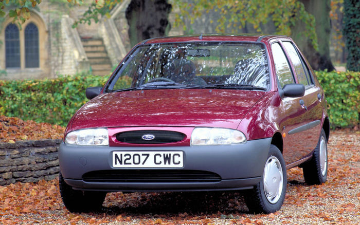 Maroon Ford Fiesta parked on fallen autumn leaves in front of a stone wall and hedge with an old building in the background. The car has a UK registration plate N207 CWC and features grey plastic bumpers and black side mirrors. This image represents a typical used car model suitable for buyers looking for reliable used cars or new cars in the affordable compact segment.