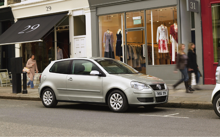 Silver Volkswagen two-door hatchback parked on a street in front of clothing shops with mannequins displaying dresses. The car has a UK license plate FN56 XYK and is positioned between shop numbers 29 and 31. Pedestrians are walking on the pavement nearby. Suitable for used cars and new cars advertising.