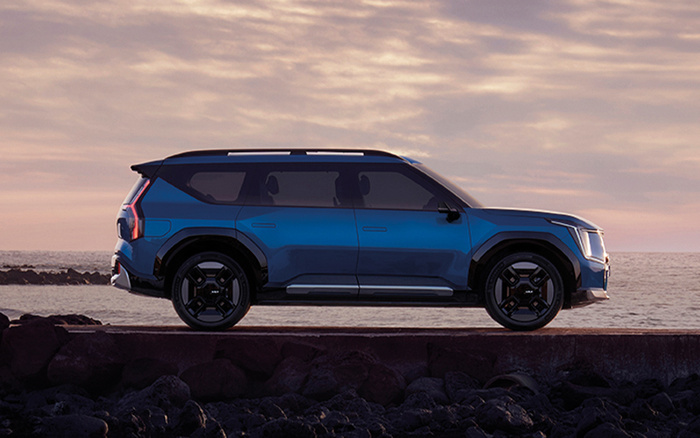 Side view of a modern blue SUV parked on a rocky shoreline at sunset with a calm sea and cloudy sky in the background, illustrating a sleek vehicle design suitable for promoting used cars and new cars.