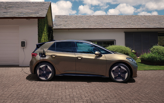 Brown electric car charging outside a modern house with a white garage door, featuring a wall-mounted charging station connected to the car. The image represents eco-friendly new cars and can be associated with used cars in the context of electric vehicle options.