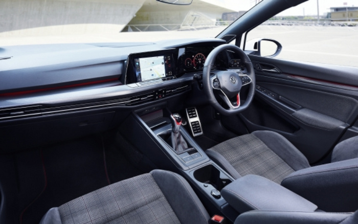 Interior view of a modern Volkswagen car featuring a digital dashboard displaying navigation and vehicle information, a multifunction steering wheel with Volkswagen logo, and front seats upholstered in a grey checkered fabric. The car is parked outdoors near a bridge, showcasing the sophisticated design and technology typical of new cars, suitable for buyers interested in used cars and new cars.