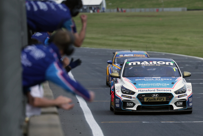 Front view of two race cars on a track, with a white and blue car in the lead displaying multiple sponsor logos including Maran Freight UK, Bristol Street Motors, Diamondbrite, Hansford Sensors, Kwik Fit, Goodyear, and S2M. Crew members in blue and white uniforms are celebrating on the left side of the image. The scene captures high-speed action on an asphalt race track with green grassy surroundings and a safety barrier in the background, highlighting the competitiveness of used cars and new cars in professional racing.