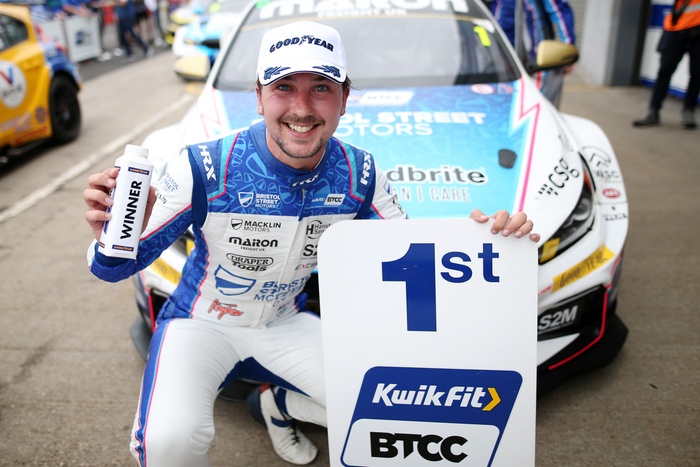 Race car driver in blue and white Bristol Street Motors racing suit and Goodyear cap holding a winner bottle and a large sign reading '1st Kwik Fit BTCC' posed in front of a race car with Bristol Street Motors branding. The image relates to new cars and used cars in the context of a British Touring Car Championship event.