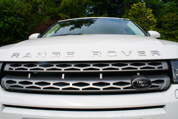 Front view of a white Range Rover with the Land Rover badge on the grille, surrounded by green trees. The image highlights the car's distinctive front grille and the Range Rover lettering on the bonnet, suitable for used cars and new cars.