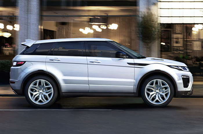 Side view of a sleek silver Range Rover Evoque driving on a city street with blurred background of buildings and lights, representing a new car option alongside used cars in an urban setting.