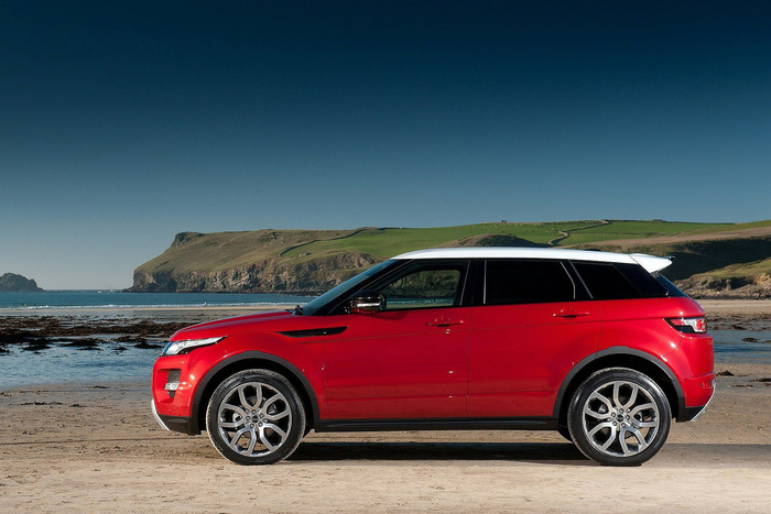 Red and white luxury compact SUV parked on a sandy beach with rocky coastline and green hills in the background under a clear blue sky, suitable for advertising used cars and new cars.