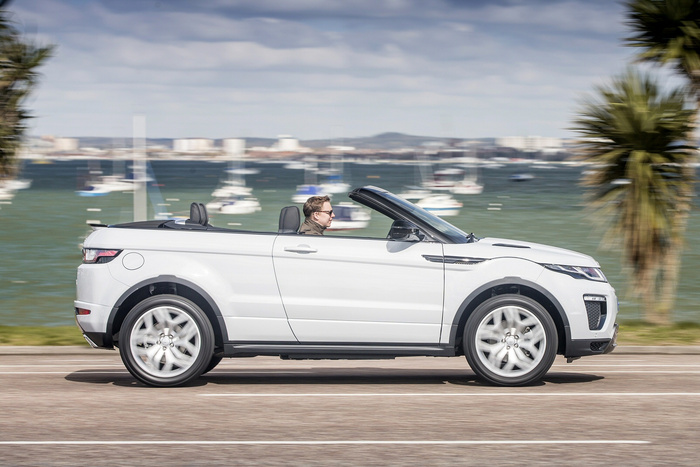 White convertible Range Rover driving on a coastal road with a man wearing sunglasses behind the wheel, palm trees and yachts in the background, representing luxury used cars and new cars for sale.