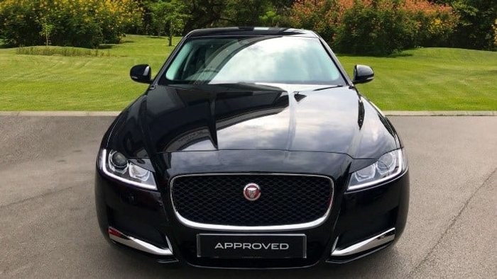 Front view of a sleek black Jaguar car with a prominent grille and logo, parked on a driveway with green grass and trees in the background. The car displays a licence plate reading 'APPROVED', indicating it as a certified vehicle, suitable for used cars or new cars buyers.