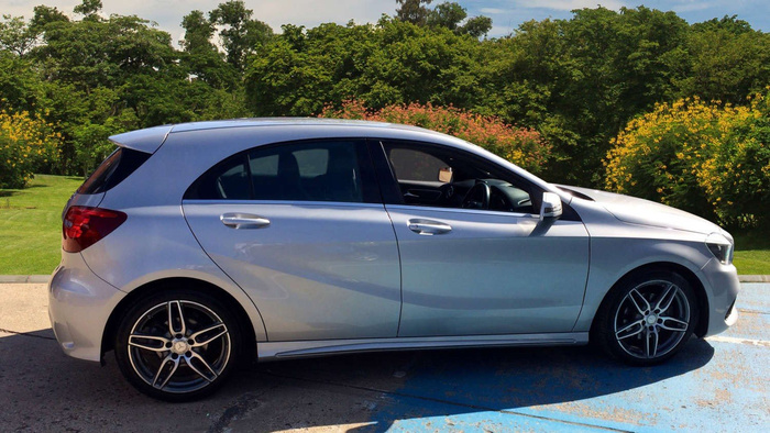 Silver hatchback car parked partially on a blue-painted disabled parking space with green trees and flowering bushes in the background, showcasing a clean side profile with tinted windows and alloy wheels, suitable for used cars or new cars sales.