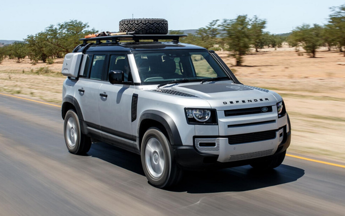 Silver Land Rover Defender driving on a paved road in a desert landscape with sparse trees. The car features a roof rack with a spare tyre and equipment mounted on it, sleek black side mirrors, and the word 'DEFENDER' prominently displayed on the front bonnet. The image showcases a rugged, off-road capable vehicle suitable for both used cars and new cars markets.