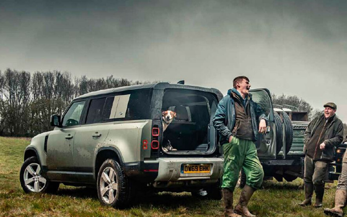 A muddy grey Land Rover Defender 110 parked on a grassy field with its rear door open, showing a dog sitting inside. Two men dressed in outdoor clothing and rubber boots stand nearby, laughing and chatting next to another vehicle. The scene suggests a countryside setting, possibly after off-road driving, highlighting rugged new cars and used cars suitable for outdoor activities.