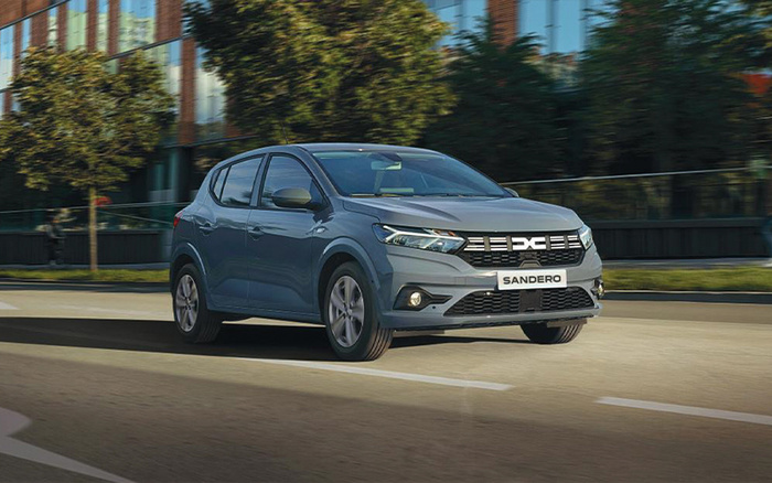 Grey Dacia Sandero hatchback car driving on a city street with modern buildings and trees in the background, showcasing a model suitable for both used cars and new cars buyers.