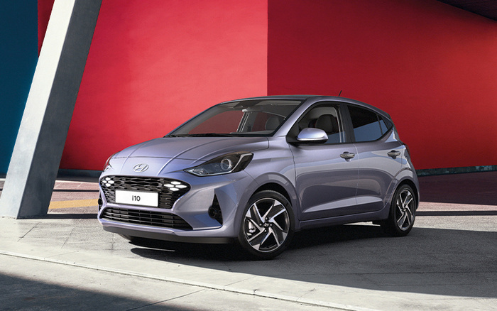 Silver Hyundai i10 hatchback car parked in front of a modern building with red and blue walls. The car is a new model, shown in a clean and well-lit environment, ideal for showcasing both used cars and new cars options.