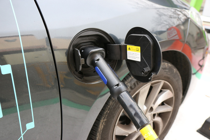 Close-up of an electric vehicle being charged, showing a black and yellow charging cable plugged into the car's charging port. The charging flap has a yellow caution label with instructions in a foreign language. The car is dark grey with blue detailing, and the wheel and part of the tyre are visible. The image represents modern new cars, including electric vehicles, highlighting the aspect of used cars transitioning to electric technology.