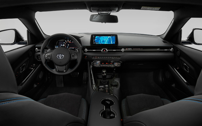 Interior view of a modern Toyota car featuring a black leather steering wheel with control buttons, digital instrument cluster, and a dashboard with a central touchscreen displaying a navigation map. The console has climate controls, gear shift, cup holders, and carbon fibre detailing. The seats are black with blue stitching, showcasing a sleek and sophisticated design ideal for buyers of used cars and new cars.