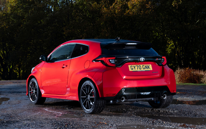 Red Toyota GR Yaris hatchback car parked on a wet gravel surface with trees in the background, showing the rear and left side of the vehicle, UK license plate GY70 GNK, highlighting sporty design and black alloy wheels, suitable for used cars and new cars listings.