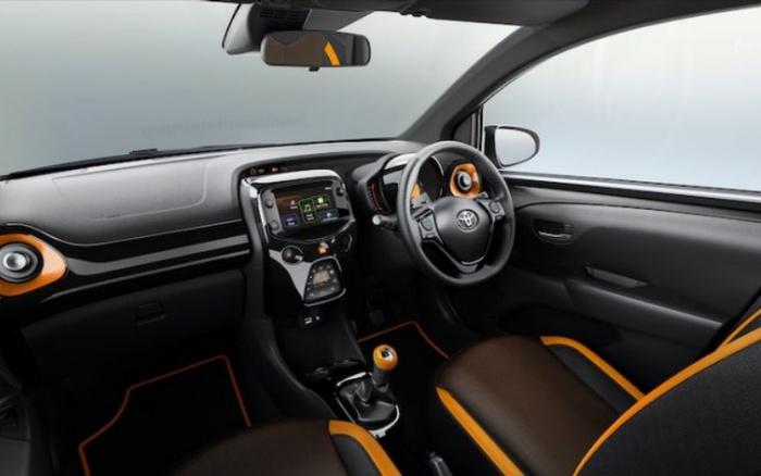 Interior view of a modern Toyota car showcasing the driver's seat, steering wheel with Toyota logo, central touchscreen display, gear shift, and black seats with orange accents. The dashboard and door panels are sleek black with orange highlights, highlighting features typically found in used cars and new cars. The image emphasises the car's tech-savvy and stylish interior design.