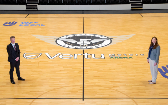 Two people standing on a basketball court at Vertu Motors Arena with Newcastle Basketball logo at centre court. The court features branding including 'Give Local' with a hand logo, and a large text reading 'Vertu Motors Arena'. The setting appears to be an indoor sports arena with seating in the background. The image highlights the association of Vertu Motors, a company dealing with used cars and new cars, with the Newcastle Basketball team.