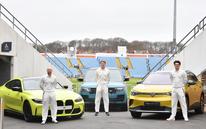 Three men in white cricket uniforms standing beside three cars parked at a sports stadium entrance. From left to right, a green BMW M4, a blue Range Rover, and a yellow Volkswagen ID.3 new cars. The stadium seating in the background is blue and yellow with advertisement boards. Signage for an accessible entrance is visible on the left. The image showcases used cars and new cars in a sports setting.