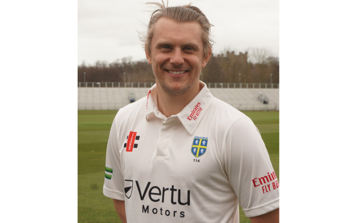 Smiling man wearing a white sports polo shirt with logos including 'Vertu Motors', 'Emirates Fly Better', 'County Championship', and a blue and yellow shield emblem with '114'. The shirt features sponsorship and branding, promoting Vertu Motors, a dealership known for used cars and new cars. The man is standing on a grass field with a stadium seating area in the background.