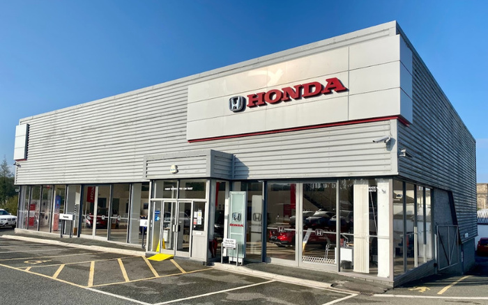 Exterior view of a Honda dealership building under clear blue sky, featuring large glass windows displaying cars inside, a prominent Honda logo and signage in red and black on a white panel above the entrance, with accessible parking spaces and a parking sign in the foreground, representing a location dealing in both used cars and new cars.