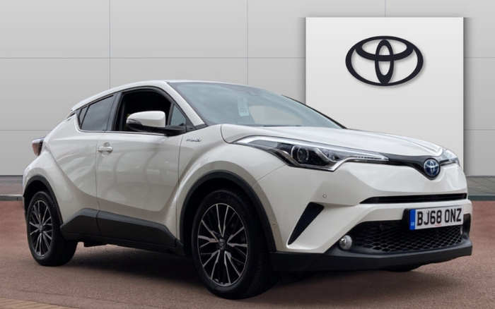 White Toyota C-HR hybrid compact SUV parked outside a Toyota dealership with a large Toyota logo on the building. The car has a UK registration plate BJ68 ONZ and features black alloy wheels and modern LED headlights. Suitable for both used cars and new cars buyers.