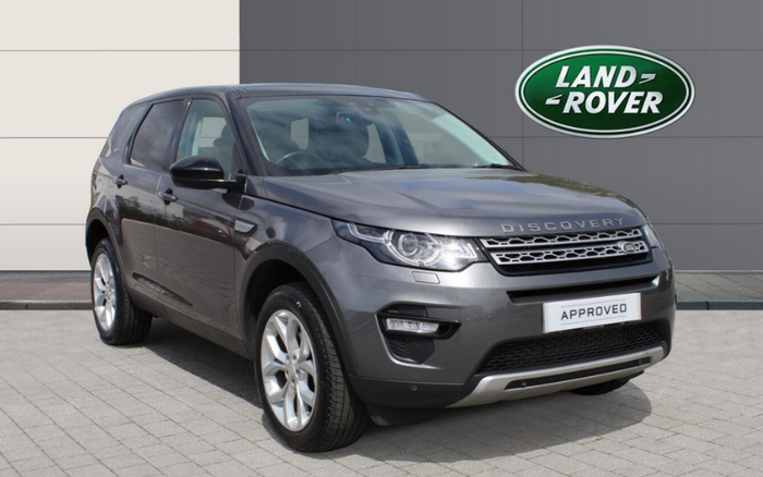Grey Land Rover Discovery parked on a paved surface in front of a grey building wall with a green Land Rover logo. The vehicle has an 'APPROVED' badge on the front grille, indicating it is a certified vehicle. This image represents used cars and new cars available from Land Rover dealerships.