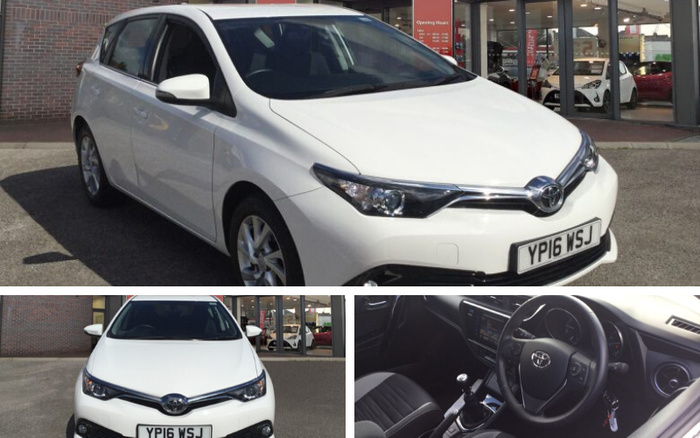 White Toyota hatchback car with registration plate YP16 WSJ parked outside a showroom displaying new cars. The showroom window shows cars inside and a red sign listing opening hours. Close-up of the car's front view and interior, featuring a black steering wheel with Toyota logo, dashboard, and manual gear shift. The image is relevant for used cars and new cars.