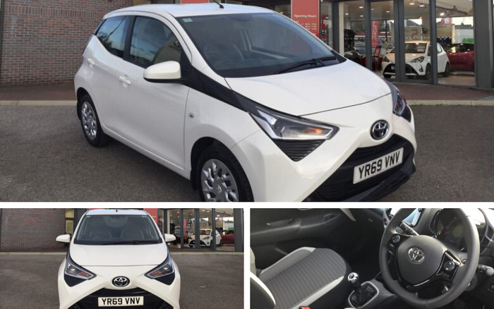 White Toyota Aygo with registration plate YR69 VNV parked outside a dealership showing the exterior front and side view, front view, and interior view of the driver's seat and steering wheel. The dealership displays various cars inside through glass windows, reflecting an environment typical for used cars and new cars sales.