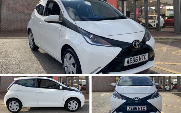 White Toyota Aygo compact car with registration plate AE66 NTC shown from front three-quarters, side profile, and front view, parked outside a dealership displaying a variety of used cars and new cars inside the showroom.