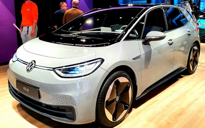 White Volkswagen ID.3 electric car displayed indoors on wooden flooring, with three people and purple walls in the background. The car features modern LED headlights and sleek design, representing a new car model ideal for customers looking for used cars or new cars.