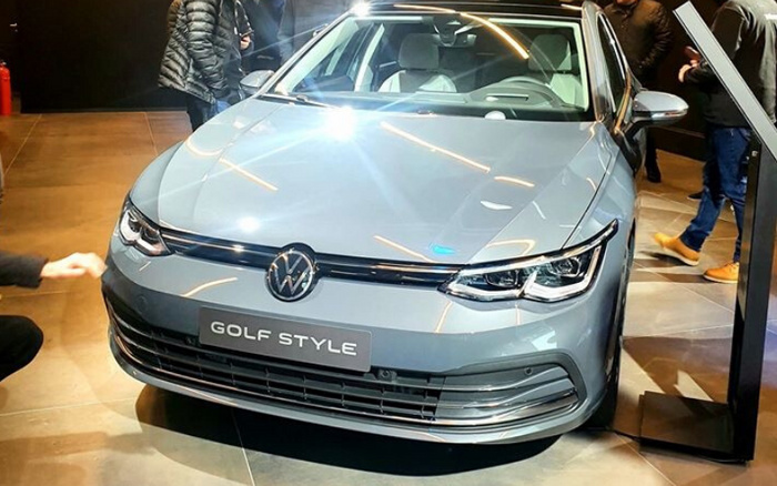 Front view of a grey Volkswagen Golf Style new car displayed indoors with shiny reflections on the hood and modern LED headlights, surrounded by people. The car is showcased on a tiled floor, highlighting the sleek design typical for new cars, relevant for used cars and new cars searches.