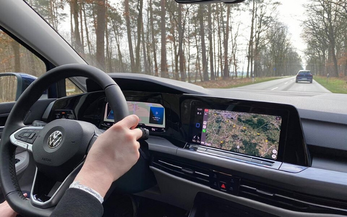 Interior view of a modern Volkswagen car dashboard with a person holding the steering wheel. The digital display behind the wheel shows navigation and driving information including a 40 mph speed limit and current speed of 94 km/h. A large central touchscreen displays a map with routes and locations. The car is driving on a two-lane road surrounded by leafless trees. This image highlights advanced digital features relevant to used cars and new cars.