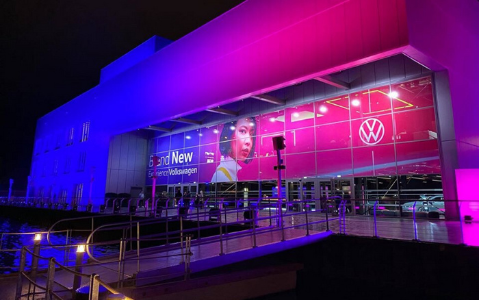 Volkswagen dealership building illuminated at night with purple and pink lighting, large glass windows displaying a banner featuring a woman's face and the text 'Brand New Experience Volkswagen'. The Volkswagen logo is prominently visible on the right side. The exterior includes a ramp and handrails with parked cars visible inside, representing both used cars and new cars available at the dealership.