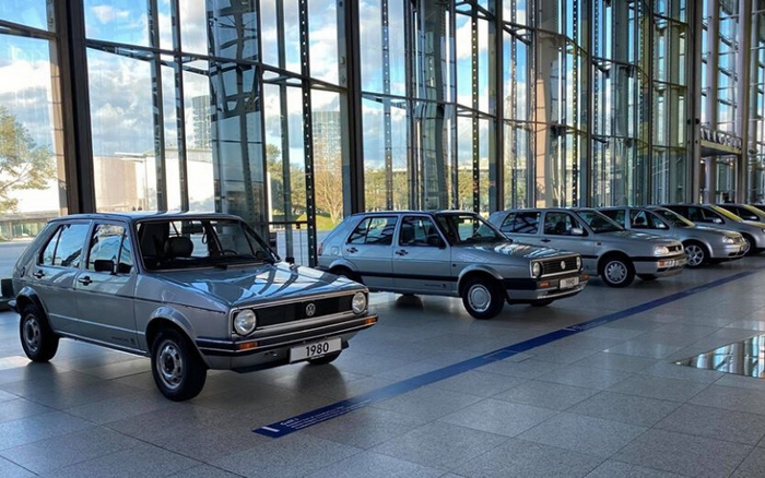 Row of Volkswagen cars displayed indoors with large glass windows in the background, showcasing models starting from a grey 1980 Volkswagen at the front, followed by similar Volkswagen cars, highlighting a timeline or evolution of vehicles. The clean, modern showroom setting emphasizes both used cars from earlier years and potentially newer models further down the line.