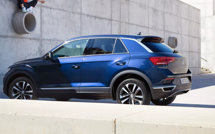 Blue Volkswagen T-Roc compact SUV parked outdoors against a modern concrete wall with cylindrical pipes, capturing the side and rear view of the vehicle with sporty alloy wheels and tinted windows, suitable for promotion of used cars or new cars.