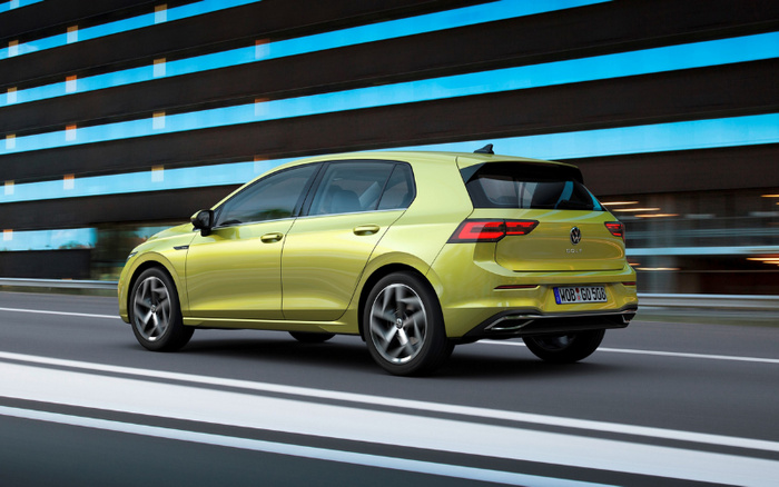 Yellow-green Volkswagen Golf hatchback driving on a road past a modern building with large windows. The car has a German license plate reading WOB GO 508. The image represents both used cars and new cars, showcasing a sleek, modern vehicle design.
