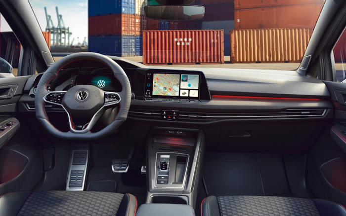 Interior view of a Volkswagen car showing steering wheel with VW logo, digital instrument cluster, and central touchscreen display featuring navigation map and media controls. The dashboard has a sleek design with red accent lighting and carbon fibre trim. Gear shifter and control buttons are visible in the central console. Outside the car, shipping containers and cranes are seen, suggesting an industrial or port setting. Suitable for illustrating used cars or new cars.