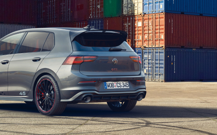Grey Volkswagen GTI hatchback parked on a concrete surface in front of stacked colourful shipping containers. The car features black alloy wheels with red accents, dual exhausts, and the license plate reads WOB CS 345. The image showcases a modern vehicle suitable for used cars or new cars market.