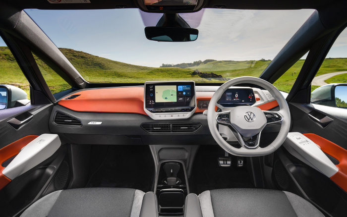 Interior view of a new Volkswagen electric car showing the dashboard and steering wheel with the VW logo. The dashboard features a digital instrument cluster displaying speed, and a central touchscreen showing a navigation map. The interior has a modern design with orange and black accents and fabric seats. The car is right-hand drive, with a clear view of green hills and a winding road outside through the windshield. This image highlights features relevant for buyers of new cars and used cars.