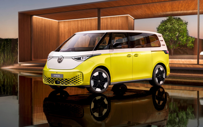 Yellow and white Volkswagen ID.Buzz electric van parked outside a modern wooden house with large glass windows at sunset. The vehicle is reflected in the shiny black surface beneath it, highlighting its sleek, futuristic design. Keywords: used cars, new cars.