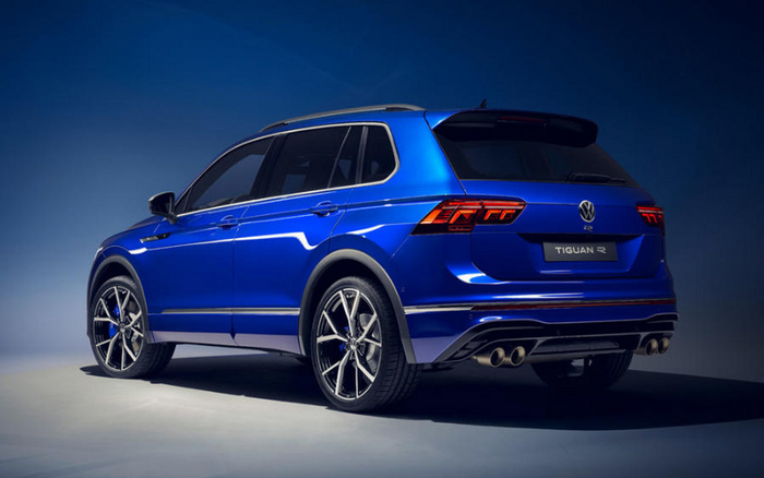 Rear side view of a blue Volkswagen Tiguan R SUV under studio lighting, showcasing its sporty design, tinted windows, dual exhaust pipes, and distinctive rear taillights. The car is positioned on a plain light grey surface with a dark blue gradient background, highlighting this model perfect for customers interested in new cars or used cars.