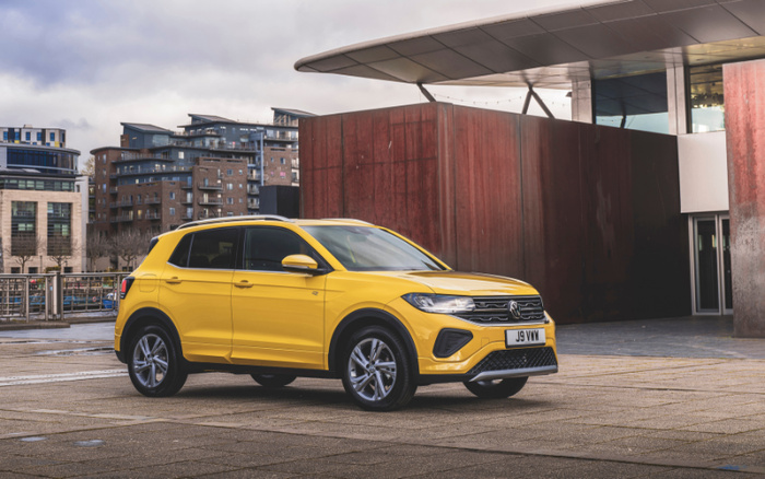 Bright yellow Volkswagen T-Cross compact SUV parked in an urban setting with modern buildings in the background. The vehicle displays a UK registration plate reading 'J9 VWW'. Ideal image for used cars or new cars listings showcasing a stylish, contemporary vehicle in a city environment.