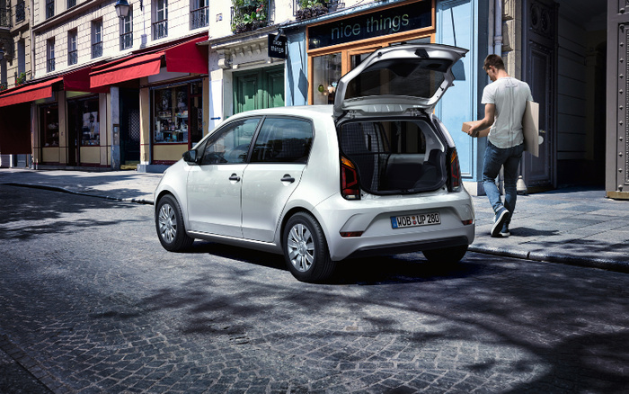 White compact hatchback car parked on a cobblestone street with its rear hatch open, showing empty trunk space. A man wearing a light grey t-shirt and blue jeans walks nearby holding a bag and a box. In the background, there is a boutique store with a sign reading 'nice things'. This urban scene highlights a new car model performance, suitable for used cars and new cars market.
